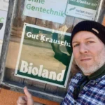 Safran Naturkost Ihr Bioladen mit Charme. Herr Sylvain Durand Inhaber und Geschäftsführer vor einem Lieferantenschild auf dem Hof von Gut Krauscha Bioland - Anbau ohne Gentechnik