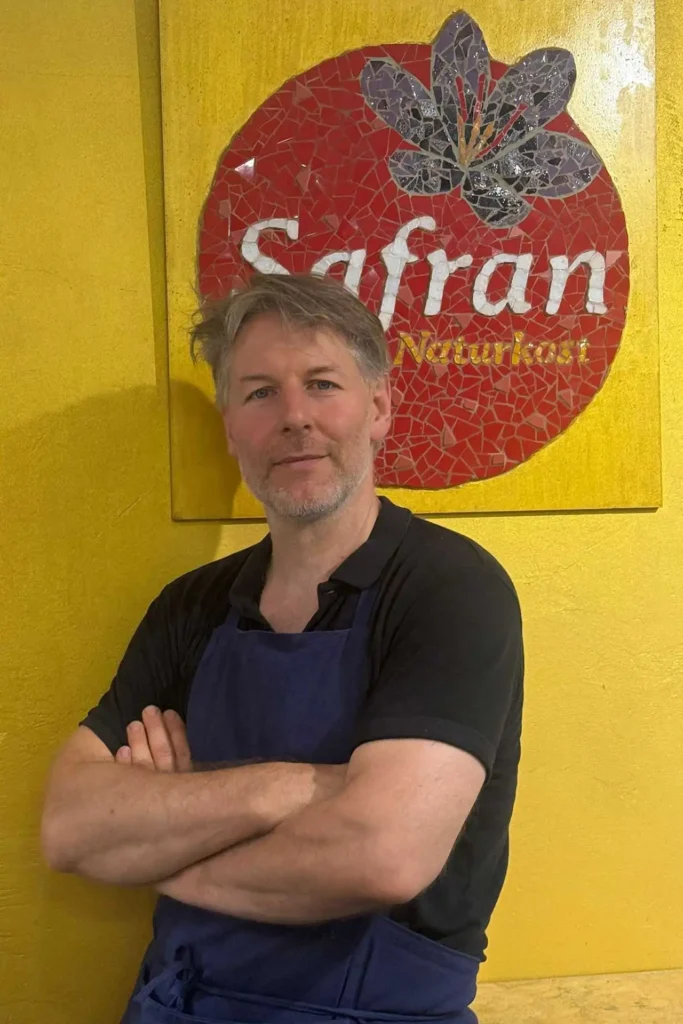 Safran Naturkost Ihr Bioladen mit Charme. Mehr als BIO. - Herr Sylvain Durand Inhaber und Geschäftsführer lachend mit verschränkten Armen zufrieden und stolz vor dem Logo im Laden