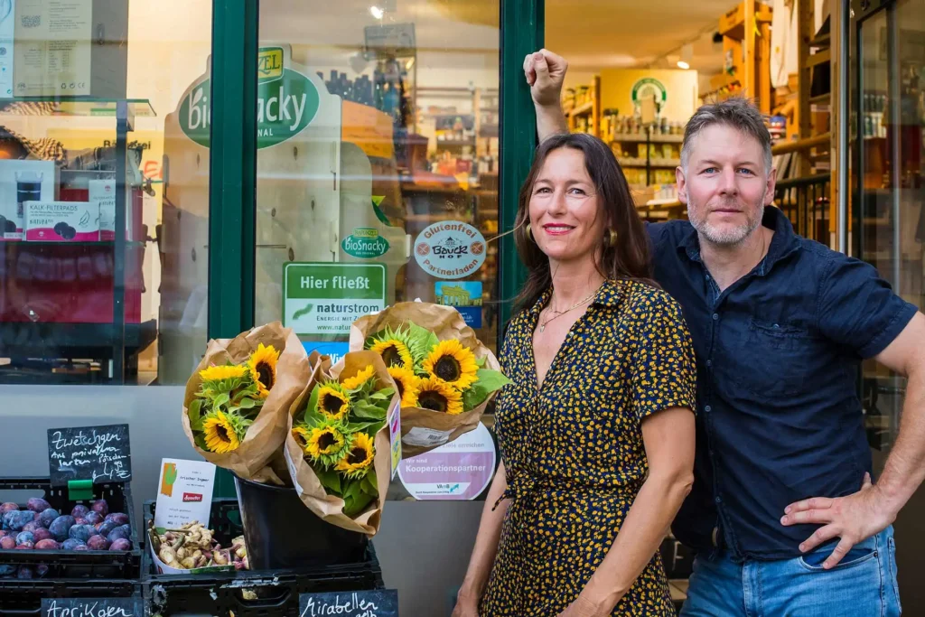 Safran Naturkost Ihr Bioladen mit Charme. Herr Sylvain Durand Inhaber und Geschäftsführer mit seiner Mitarbeiterin vor dem Schaufenster des Ladens - frisches Obst, Genüse und Sonnenblumen