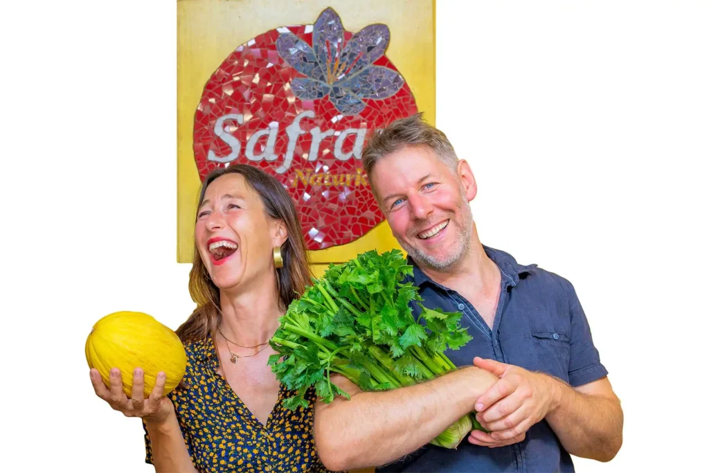 Safran Naturkost Ihr Bioladen mit Charme. Herr Sylvain Durand Inhaber und Geschäftsführer mit seiner Mitarbeiterin lachend vor dem Logo im Laden Honigmelone in der Hand - Sellerie im Arm
